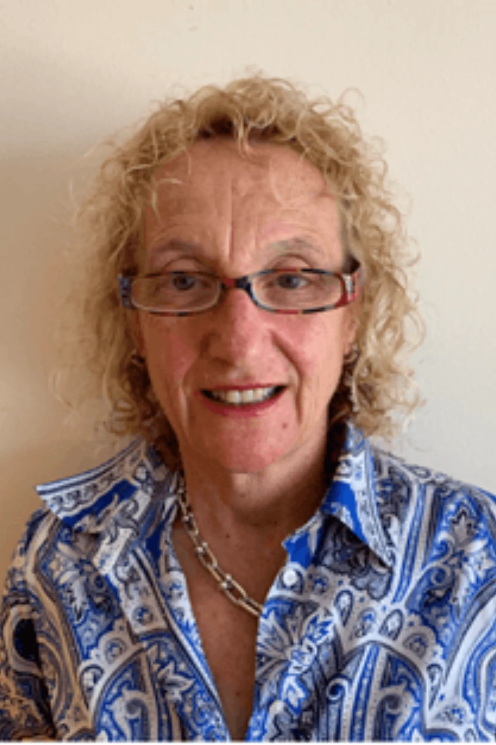 Smiling older woman with curly blonde hair, glasses, blue patterned shirt, and a silver necklace against a light background